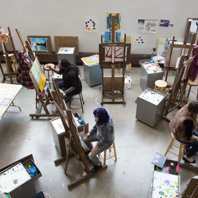 In an art studio, students are seated in front of easels, drawing or painting.