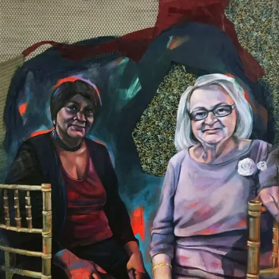 Two older women sitting in chairs, the one in front  in a purple dress and the one in the back a dark red dress.