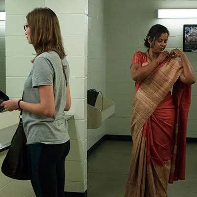 Two women in a public restroom. One is holding a baseball cap and looking in the mirror; the other is adjusting her sari.