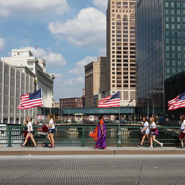 Nirmal Raja and Lois Bielefeld, Downtown – Sari 18 (flags), 2017, archival pigment print, 44 x 60” Pedestrians crossing a bridge over a river in an urban setting. Most are probably part of a group. They're dressed similarly (in white tops and blue shorts or pants) and are all walking in the same direction. A woman in a pink and purple sari is walking in the opposite direction.