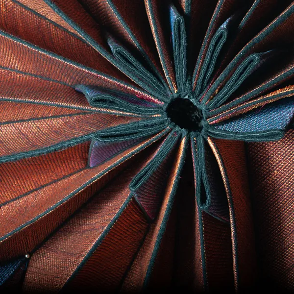 Close-up of copper-colored accordion-pleated fabric curled in a circle. Each pleat is edged in black, and is notched with a black pleat at one edge.