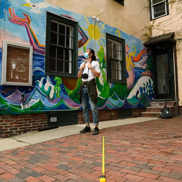 A woman standing outside in front of a mural of the sea and a sunny sky and skeletal arms waving in the air. A tape measure on the ground measures the distance between the photographer and the subject.