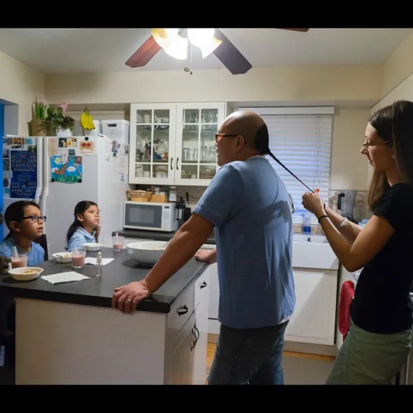 Mike Huie standing in a kitchen. A woman stands behind him, braiding his hair.Three children face them from the other side of a kitchen island.