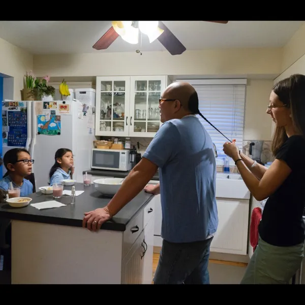 Wing Young Huie, He called himself Mike, 2016, Digital Light Jet print, 20 x 30” Mike Huie standing in a kitchen. A woman stands behind him, braiding his hair.Three children face them from the other side of a kitchen island.