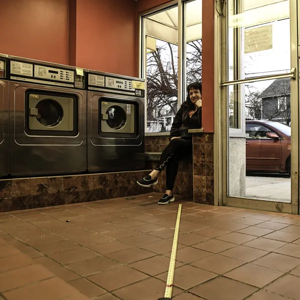 Woman sitting in a laundromat. A tape measure on the ground measures the distance between the photographer and the subject.
