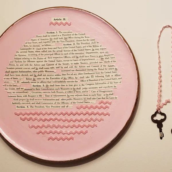 A pink plate with strips of paper glued on it that contain (parts of) Article II, Sections 1-4 of the United States Constitution. There's also some pink bric-a-brac on the plate. To the right of the plate is pink bric-a-brac with an old-fashioned key on each end.