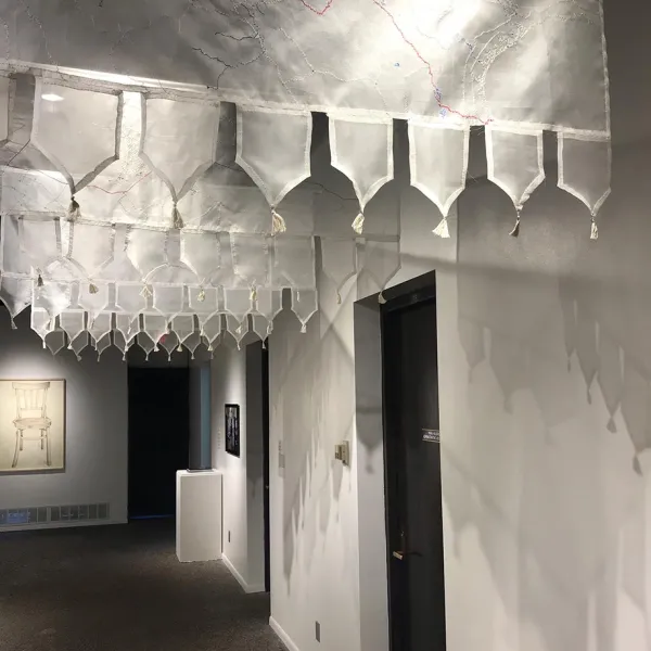 Nirmal Raja, Fault Lines: North Korea/South Korea, India/Pakistan, Syria/Turkey, Gaza Strip, United States/Mexico, 2019, hand embroidery on silk organza, clear acrylic dowels, 5 panels, each 21 x 48” Five gauzy hangings with the "fault lines" embroidered on them, hanging over two black doors on the right wall of a white hallway.