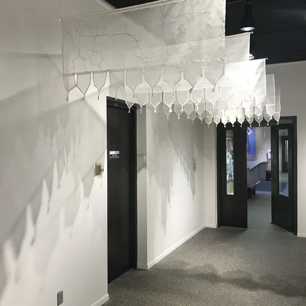 Five gauzy hangings with the "fault lines" embroidered on them, hanging over a black door on the left wall of a white hallway.