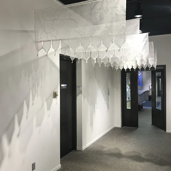 Nirmal Raja, Fault Lines: North Korea/South Korea, India/Pakistan, Syria/Turkey, Gaza Strip, United States/Mexico, 2019, hand embroidery on silk organza, clear acrylic dowels, 5 panels, each 21 x 48” Five gauzy hangings with the "fault lines" embroidered on them, hanging over a black door on the left wall of a white hallway.