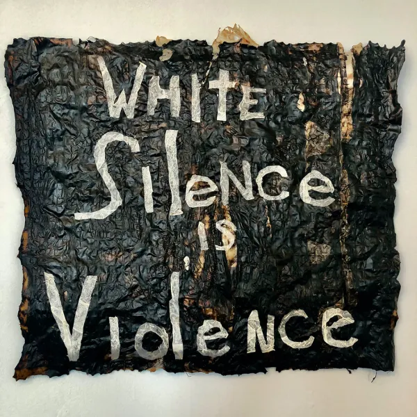 Carolyn Halliday, "Skin in the Game," animal membrane dyed with black walnut, used dryer sheets, commercial felt, thread, 22” x 22" The words "WHite Silence is Violence" in white against a background of animal membrane dyed black.