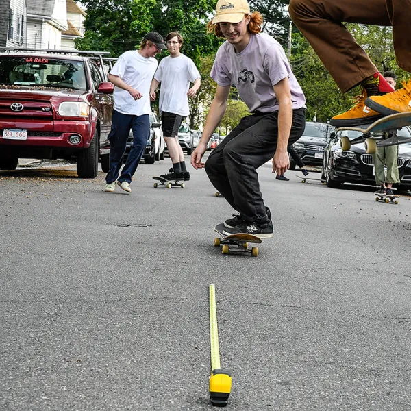 Teenagers skateboarding in a parking lot. A tape measure on the ground measures the distance between the photographer and the closest skater.