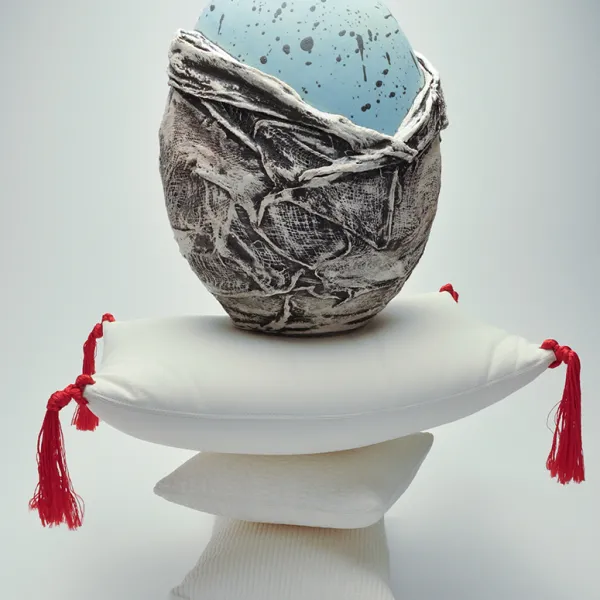 Mika Negishi Laidlaw, Remember, 2019, slip cast porcelain and red thread, 19 x 13 x 10" A speckled, light blue egg, two-thirds of which is covered by gray and white material. This is on top of a white pillow with four red tassels, and that pillow is balanced on top of two smaller white pillows.