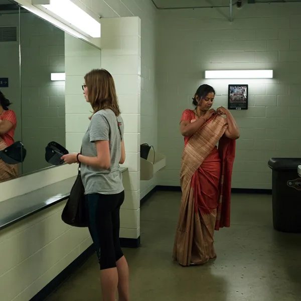 Two women in a public restroom. One is holding a baseball cap and looking in the mirror; the other is adjusting her sari.