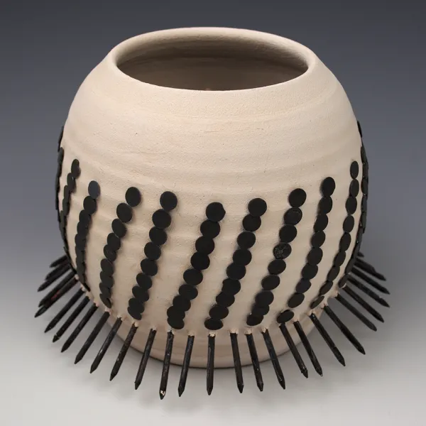 Large round beige ceramic bowl with repeating diagonal (almost vertical) rows of black circles. There are nails painted black sticking out all around the bottom of the bowl.