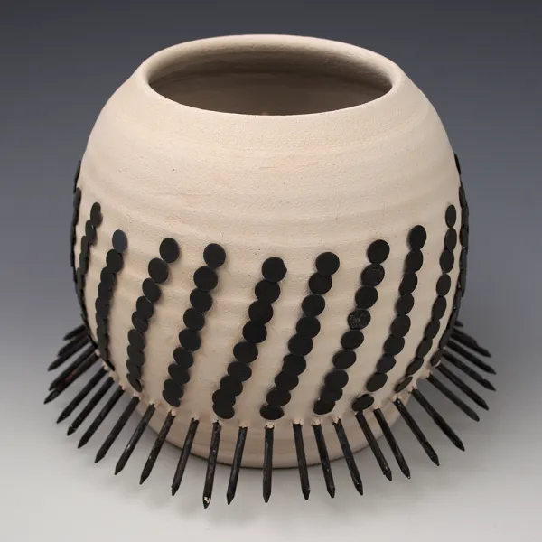 Tavia VanZuilen, Two Hundred Thirty Three, 2016, raku, nails, 6.5 x 8.5 x 8.5” Large round beige ceramic bowl with repeating diagonal (almost vertical) rows of black circles. There are nails painted black sticking out all around the bottom of the bowl.