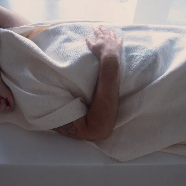 A person laying on their back, wrapped in a white blanket, with their arm over their head