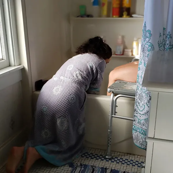A woman on her knees bending over to wash a person sitting on a chair in a shower
