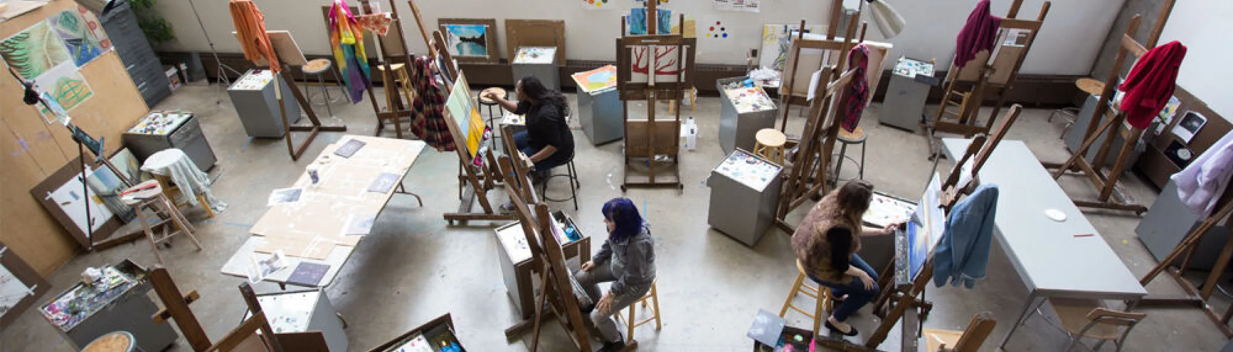 In an art studio, students are seated in front of easels, drawing or painting.