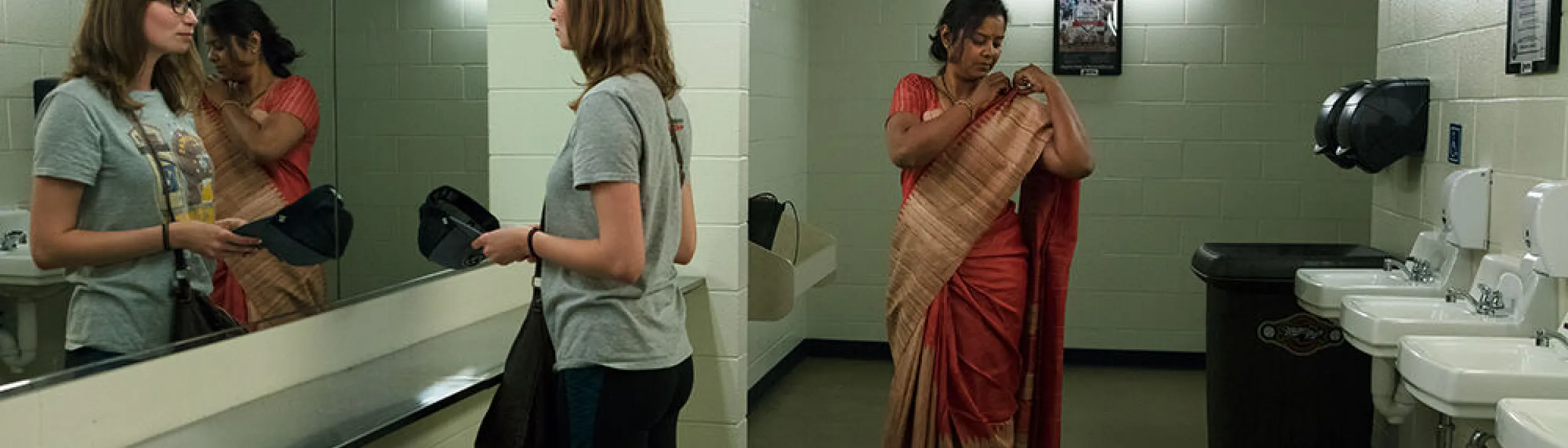 Two women in a public restroom. One is holding a baseball cap and looking in the mirror; the other is adjusting her sari.