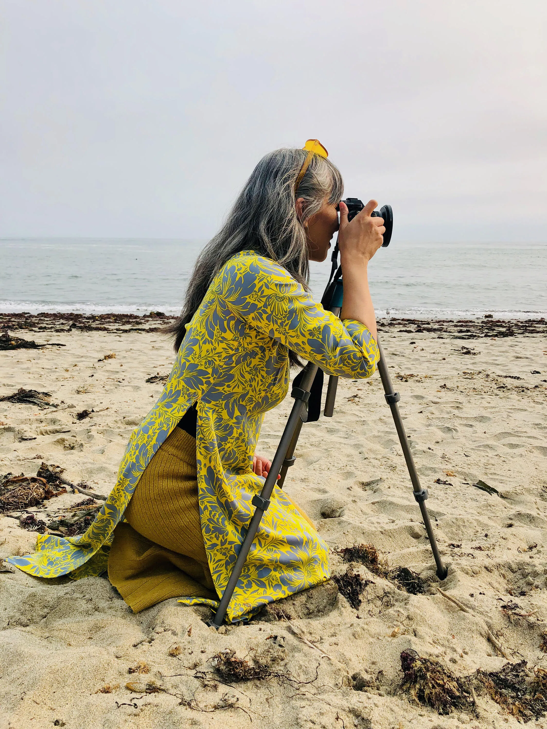 A woman in a yellow dress kneeling and taking a photograph on the beach.