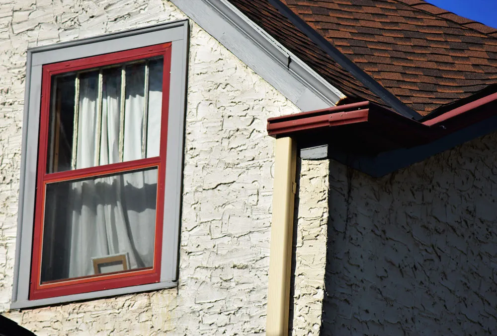 Roofline corner of stucco house, one window outlined in read, brown shingles