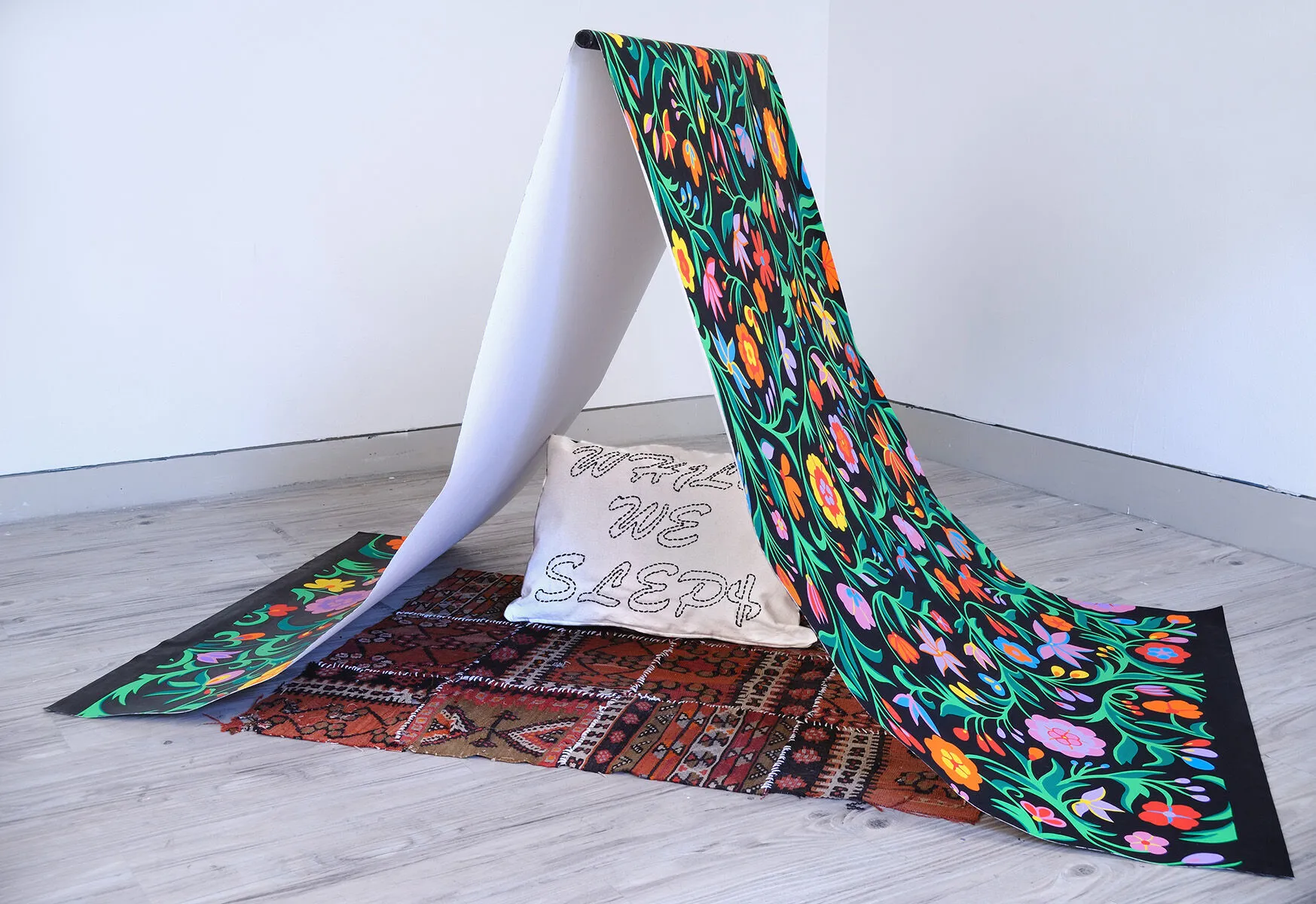 "HomeLoss" - A canvas painted with a colorful acrylic pattern on cavas; it is held up like a tent, under it is a Persian rug and a pillow that reads "we sleep here"