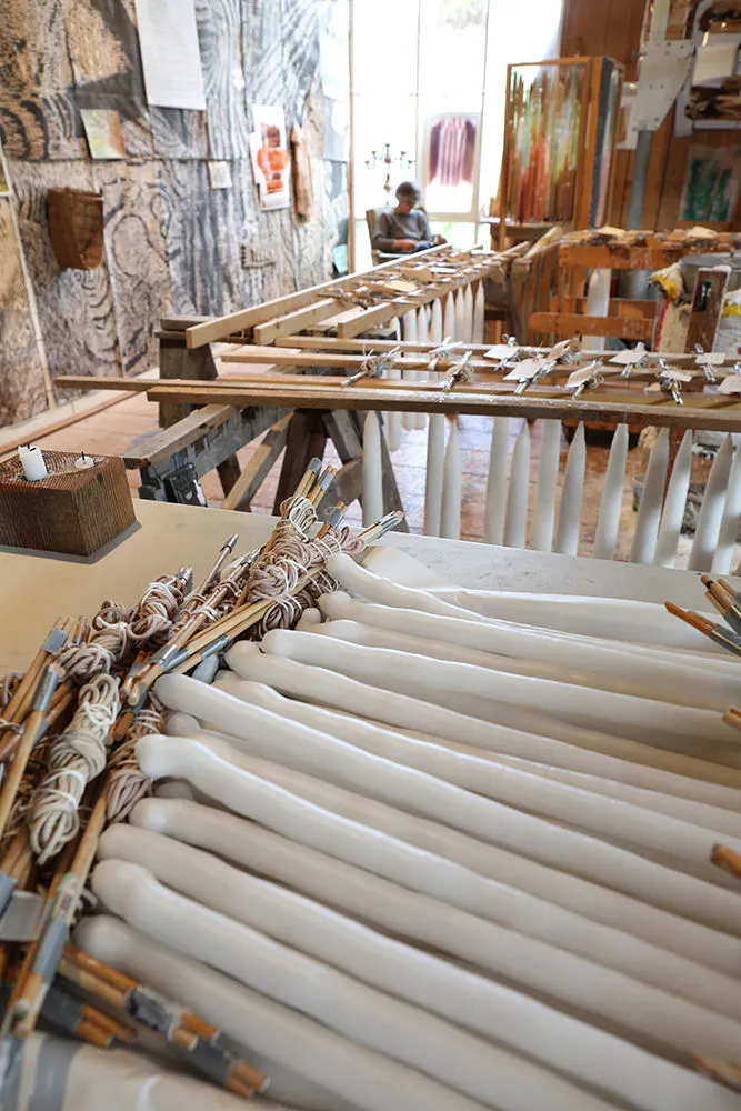 The artist sits in a distant corner of an art studio full of candles being on tables and hanging to dry on racks.