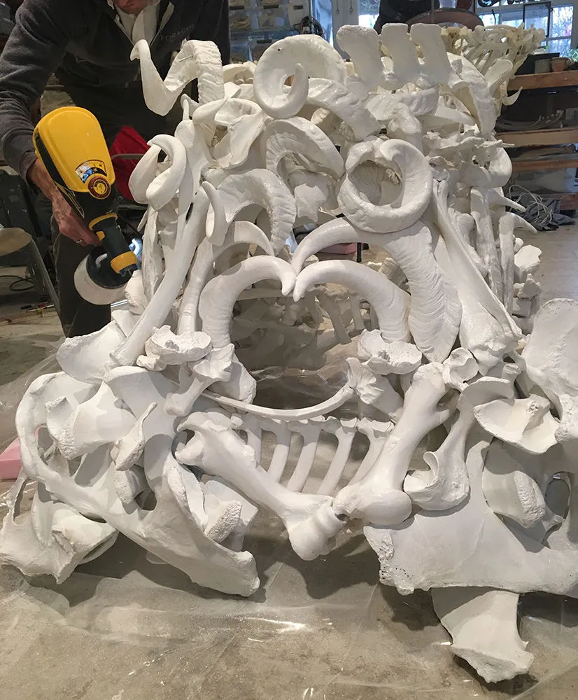 Judy Onofrio in her studio working with a power tool on a sculpture  of a pile of large bones and tusks.
