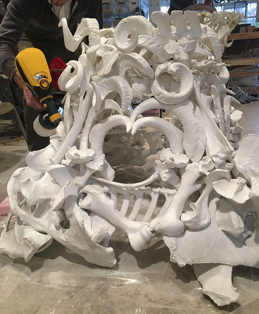 Judy Onofrio in her studio working with a power tool on a sculpture of a pile of large bones and tusks.